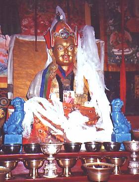 With a steady gaze, Padmasambhava’s right hand holds an upright gold vajra to his heart. His left hand is placed in his lap and holds a white skull cup filled with nectar, jewels, and a long-life vase